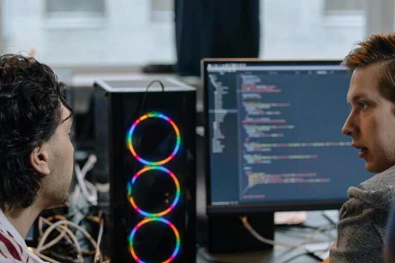 Two programmers discussing code on a monitor in a tech workspace, focusing on collaboration.