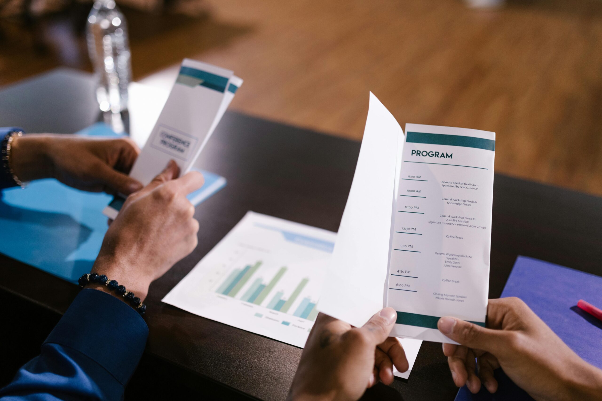 Hands examine program guide in a corporate meeting setting, highlighting agenda details.